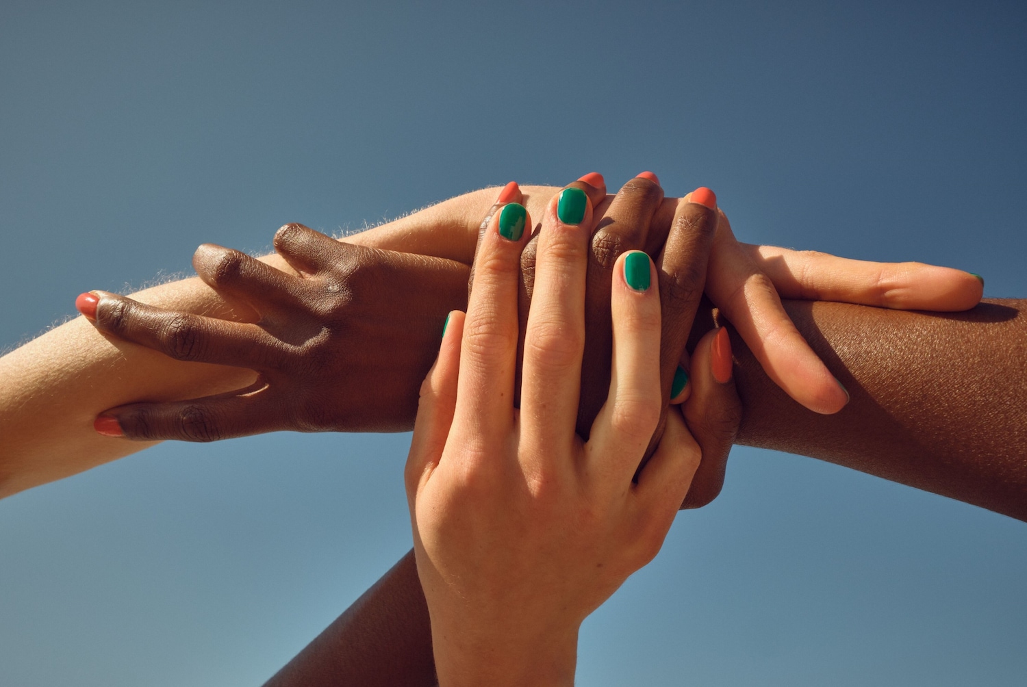 Gros plan sur des mains de différentes couleurs de peau, les ongles vernis en vert et orange, entrelacées sur un fond bleu ciel.
