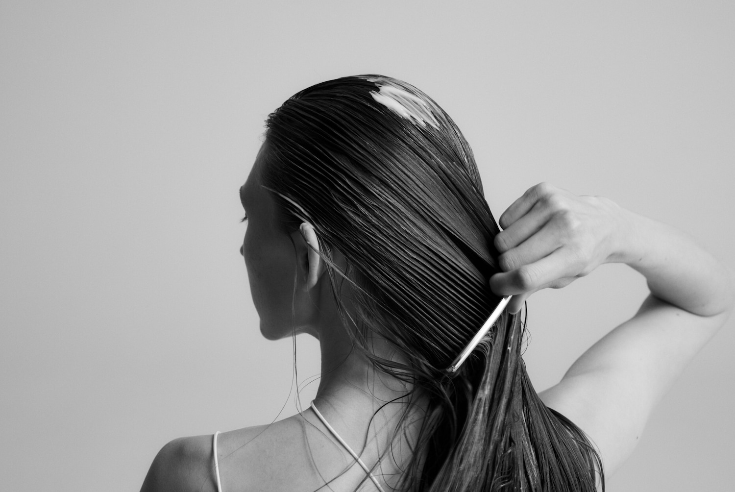 En noir et blanc, une femme démêle ses longs cheveux mouillés avec un peigne, vue de dos, mettant en valeur le soin capillaire appliqué.