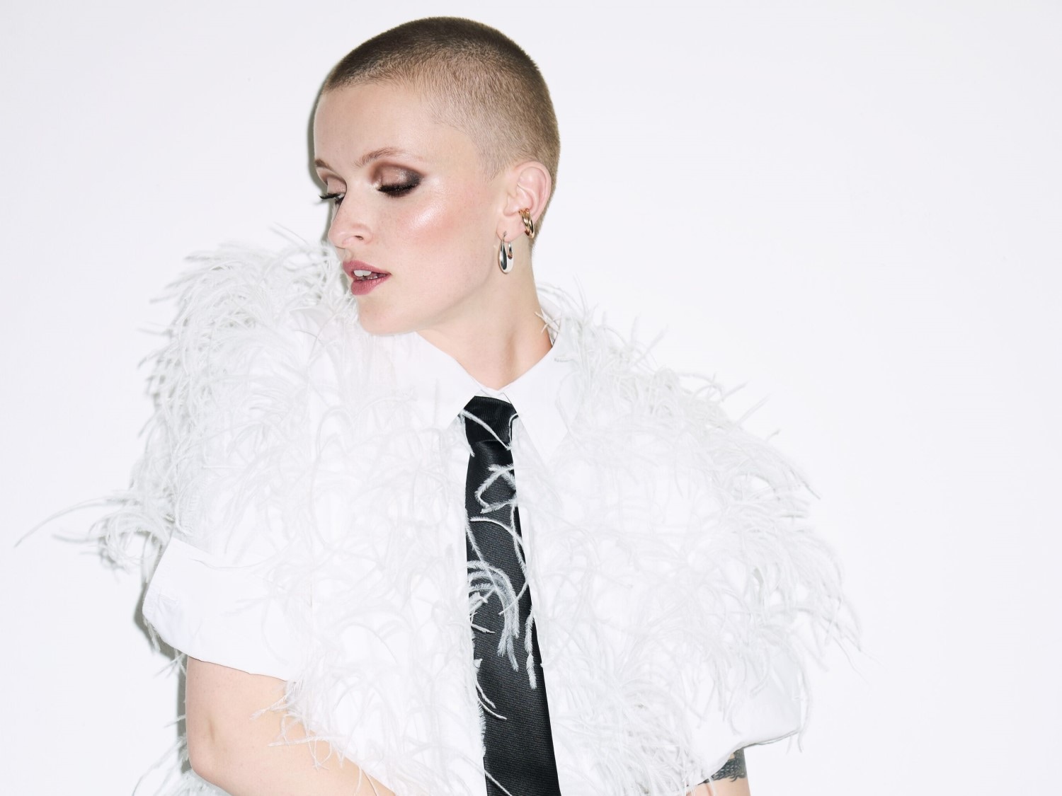 Porträt einer Frau mit rasiertem Kopf, die ein weißes Hemd mit Federn und eine schwarze Krawatte trägt, während sie zur Seite schaut.