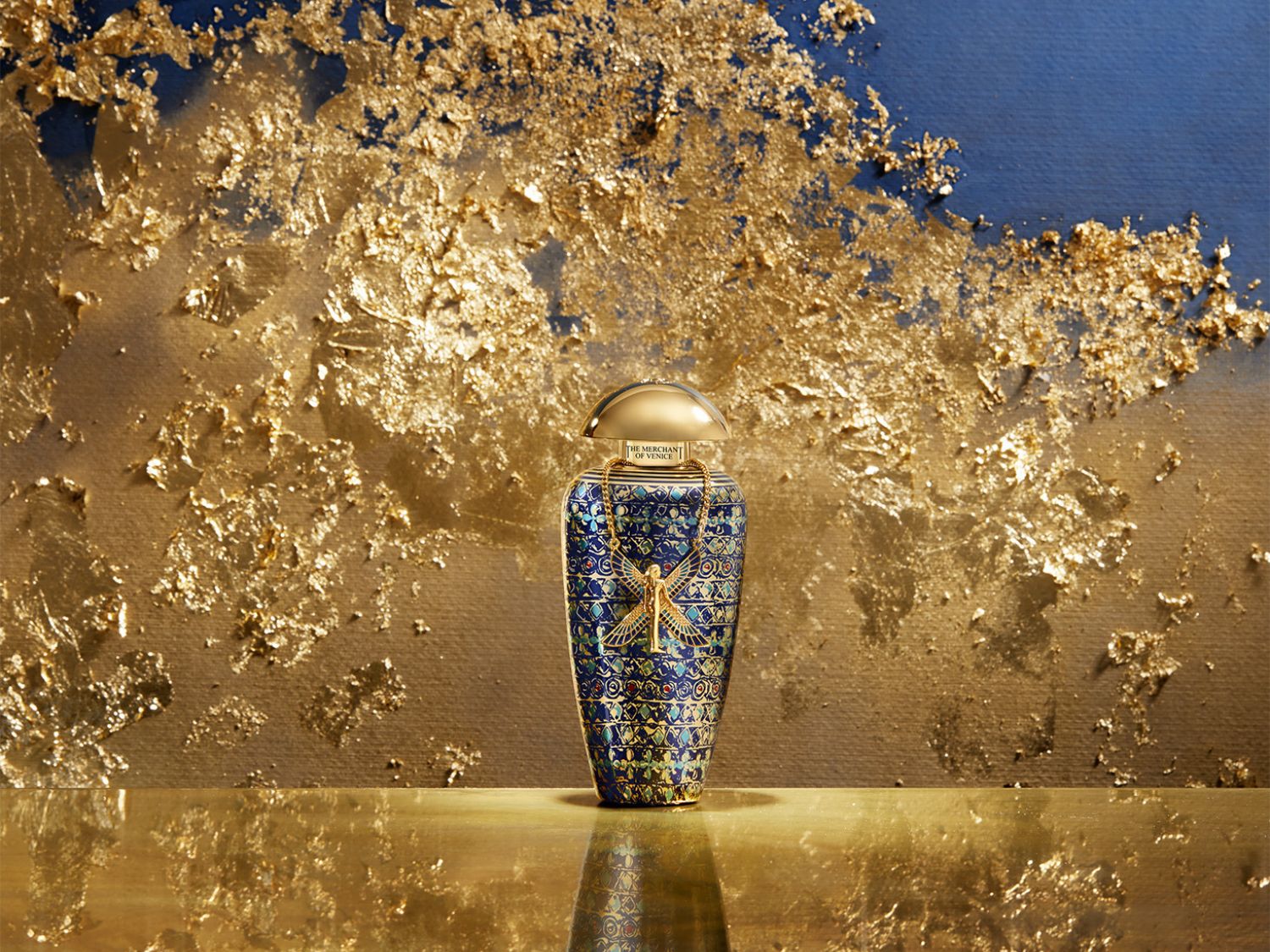Lahvička parfému Merchant of Venice má modrý vzorovaný povrch se zlatým víčkem a zlatým přívěskem ve tvaru anděla, vše na pozadí zlaté textury.