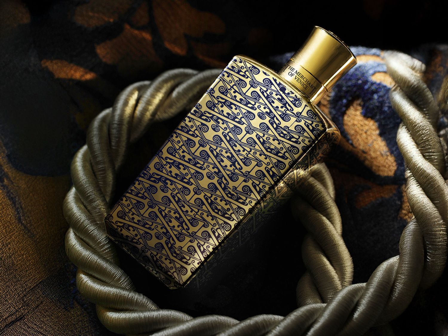 Zlatá obdélníková lahvička parfému s modrými ornamenty a zlatým uzávěrem, ležící v ozdobném kruhu.
