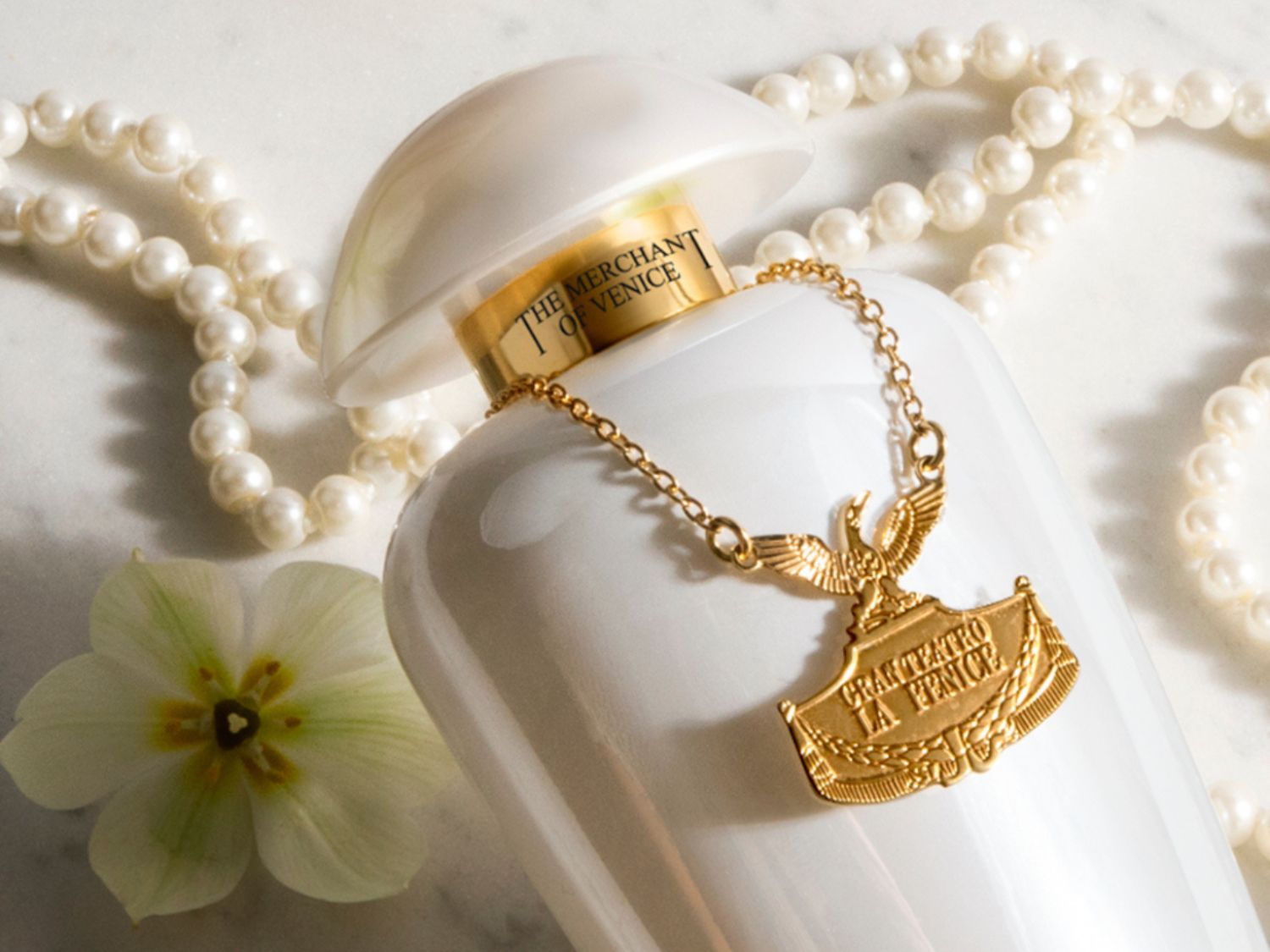 Bílý flakon parfému The Merchant of Venice zdobí zlatý přívěsek a perlový náhrdelník, doplněný jemným květem, vytvářející luxusní a elegantní kompozici.