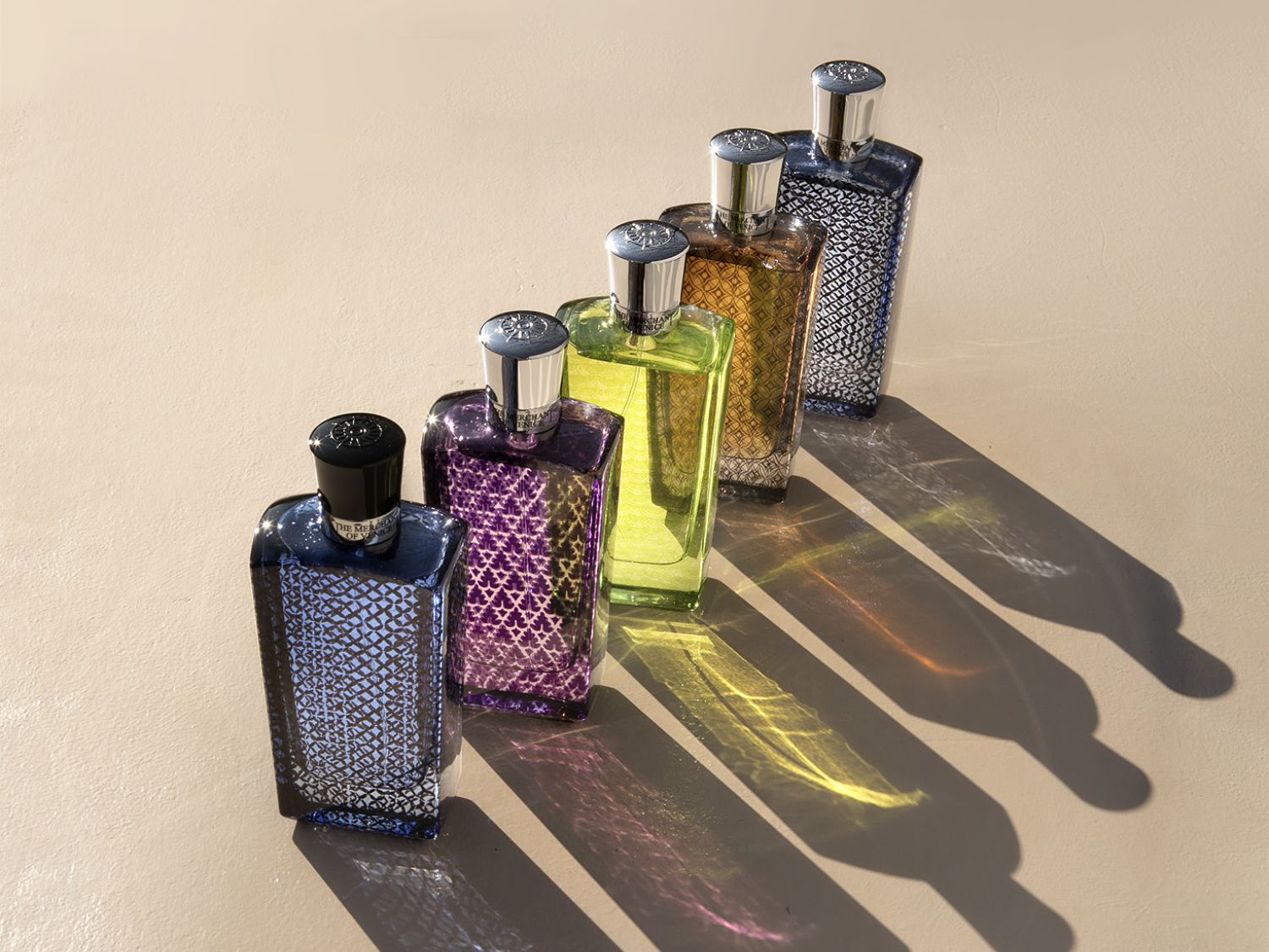 Pětice flakonů parfémů v různých barvách, od modré přes fialovou, zelenou, hnědou až po stříbrnou, stojí v řadě a vrhají dlouhé stíny na béžovém pozadí.