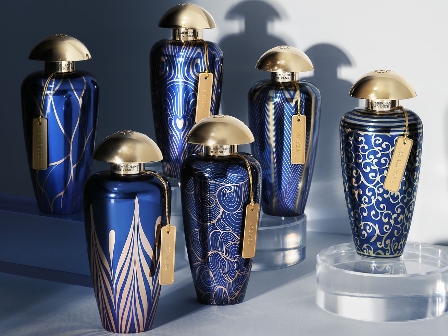 Šest modrých parfémových flakonů Merchant of Venice s různými zlatými vzory a zlatými víčky ve tvaru kupole.