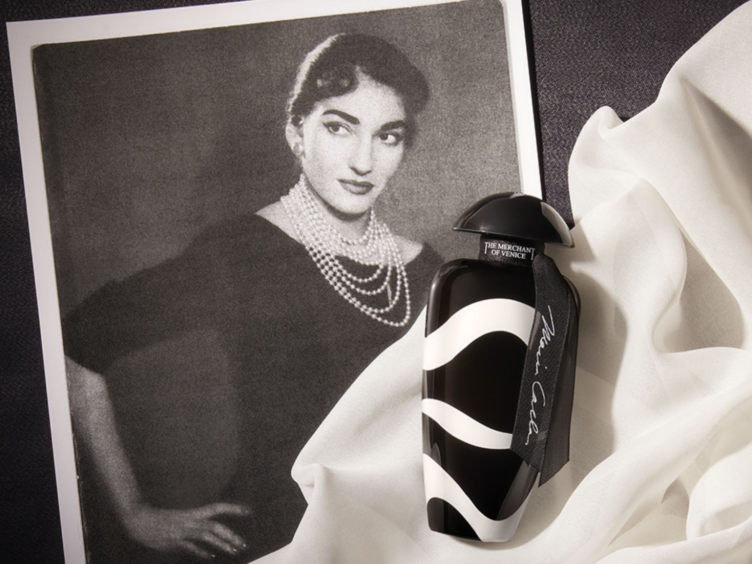 Černobílá fotografie Marie Callas vedle flakonu parfému The Merchant of Venice s černobílým vlnitým designem a černou stuhou.