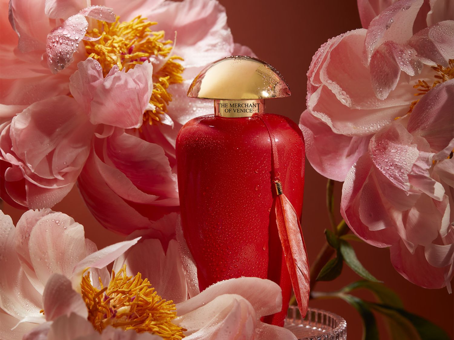 Červený flakon parfému The Merchant of Venice s zlatým víčkem, obklopený růžovými pivoňkami s kapkami vody, vytváří luxusní a svěží dojem.