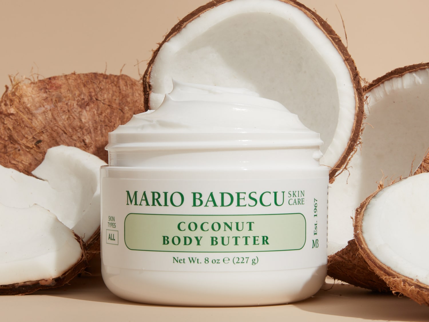 Een pot Mario Badescu Coconut Body Butter, omringd door kokosnoten, belooft een hydraterende ervaring voor alle huidtypes.