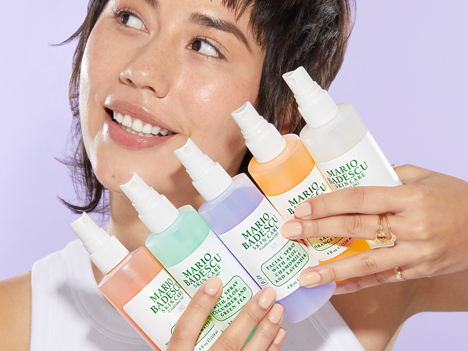 Een stralende vrouw toont een collectie Mario Badescu gezichtssprays in pastelkleurige flessen, elk met een unieke formule voor een verfrissende huidverzorgingsroutine.