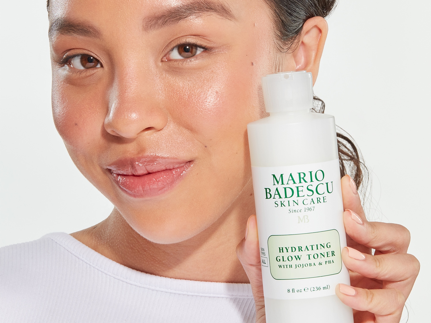Een vrouw houdt een witte fles Mario Badescu Hydrating Glow Toner vast, met een groen etiket dat de formule met Jojoba & PHA benadrukt, voor een stralende huid.