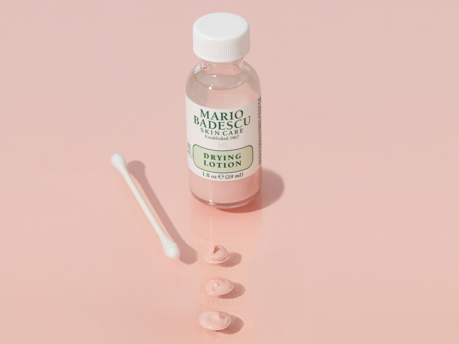Mario Badescu Skin Care Drying Lotion in een heldere fles met een witte dop, naast een wattenstaafje en drie kleine roze lotiondruppels.