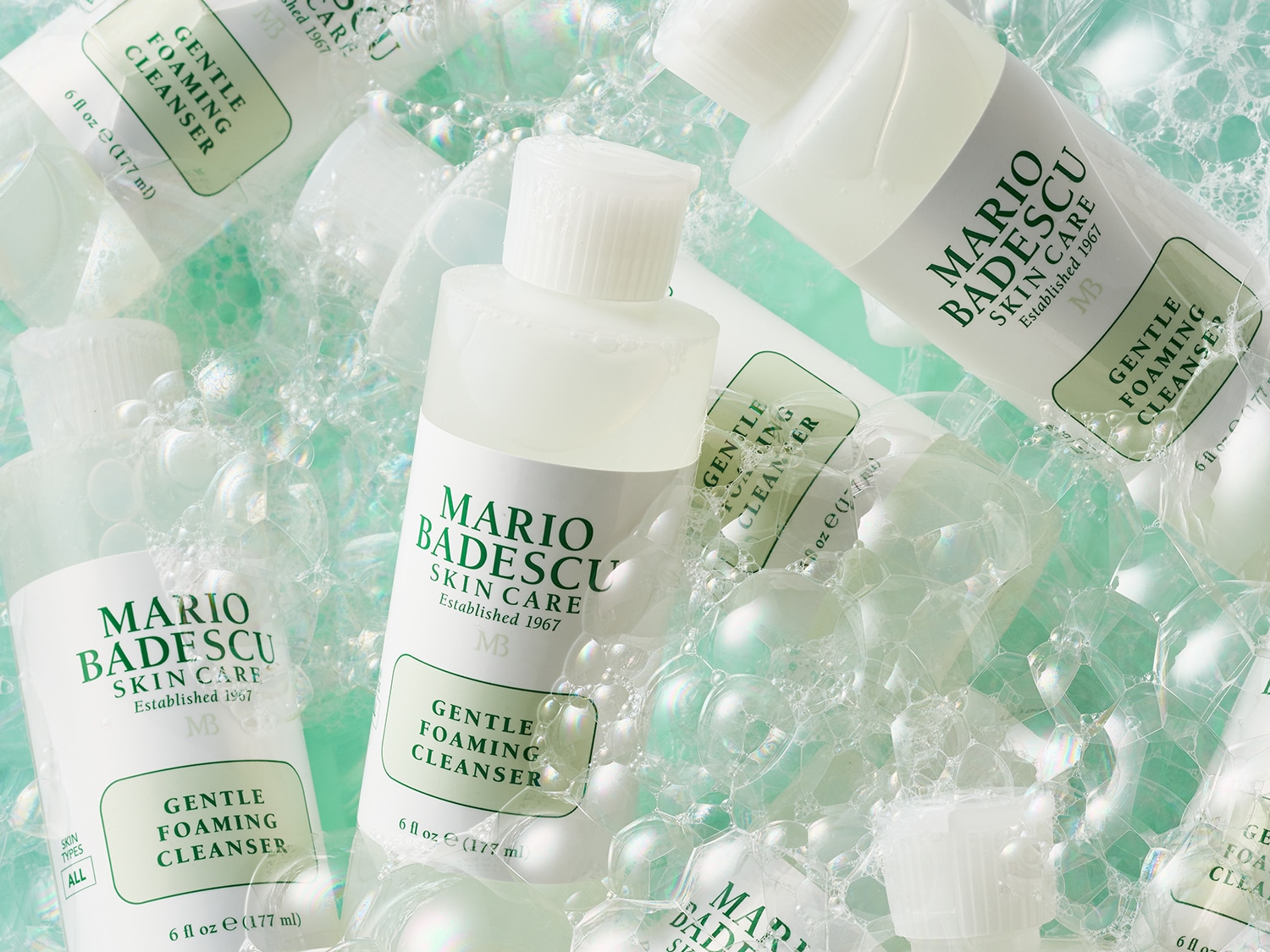 Verschillende flessen Mario Badescu Gentle Foaming Cleanser omgeven door bubbels, met de tekst 'Mario Badescu Skin Care Established 1967 MB Gentle Foaming Cleanser 6 fl oz e (177 ml)'.