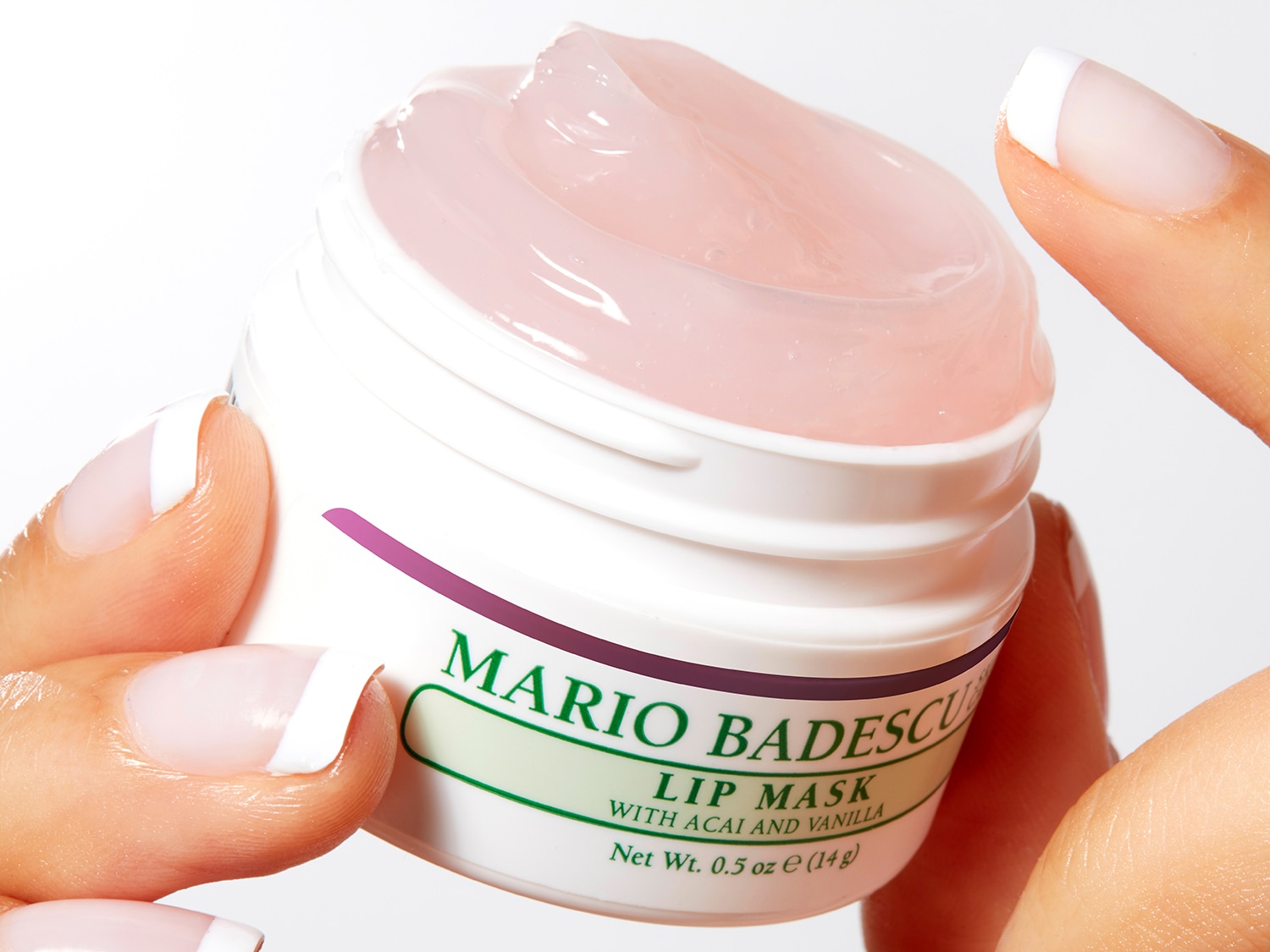 Een close-up van een witte pot Mario Badescu Lip Masker met acai en vanille, de pot is gevuld met een roze gel en wordt vastgehouden door een persoon met een witte manicure.