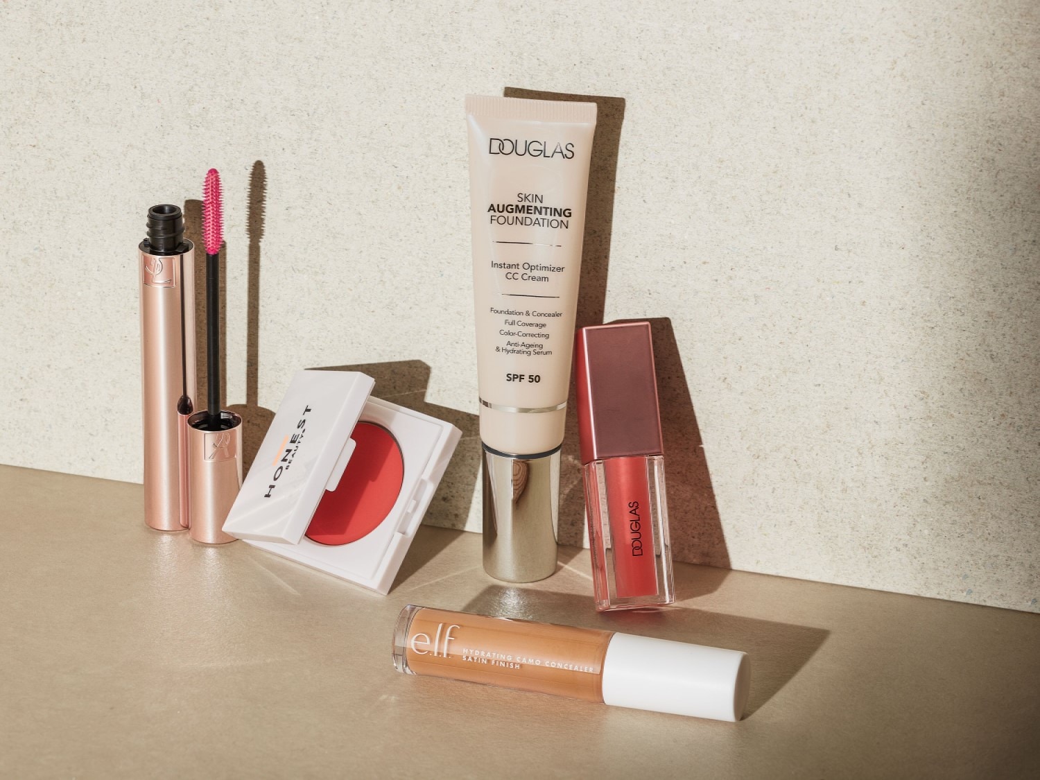 Assortment of beauty products including Douglas Skin Augmenting Foundation, Honest Beauty blush, e.l.f. Hydrating Camo Concealer, and Douglas lip gloss.