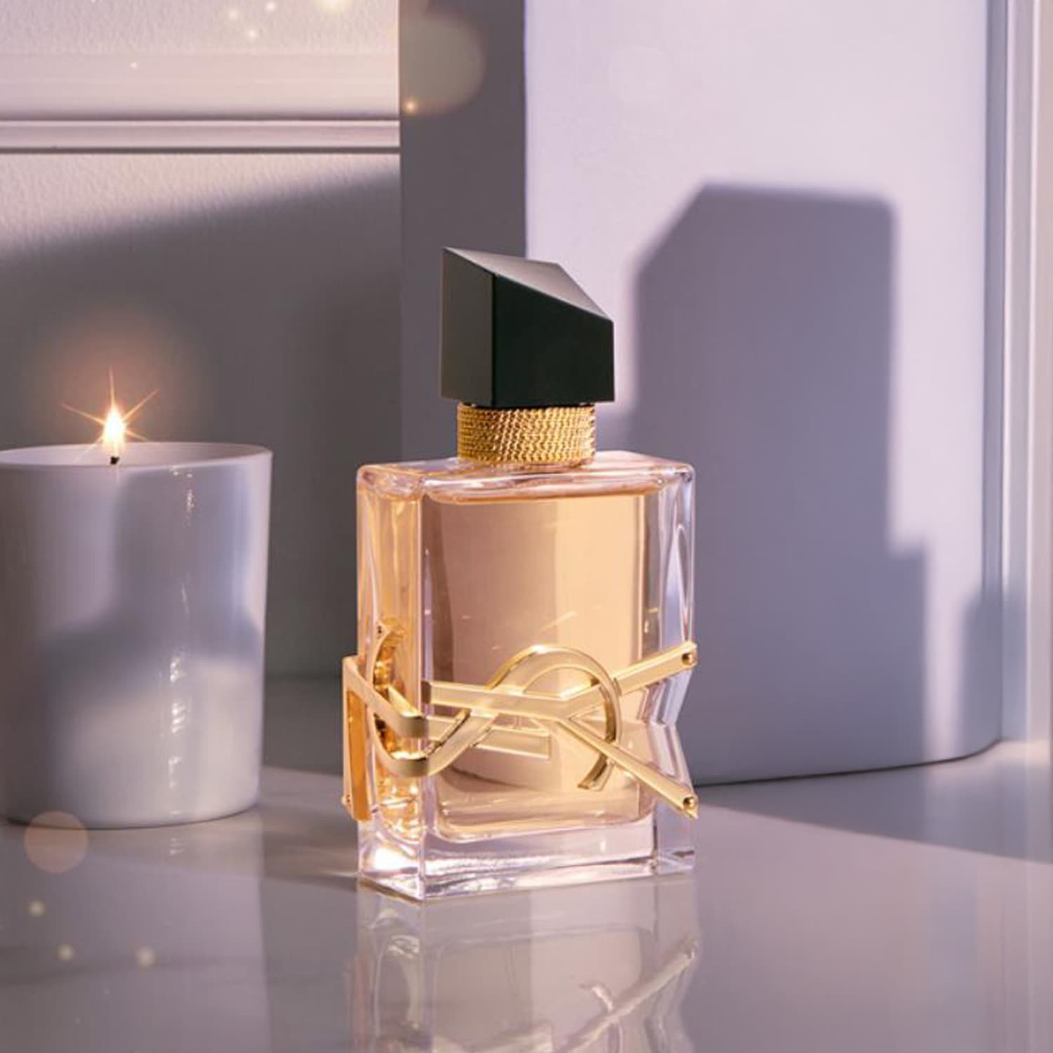 Perfumy YSL Libre w przezroczystej butelce ze złotym logo YSL, czarnym korkiem i złotą szyjką, obok zapalonej świecy.