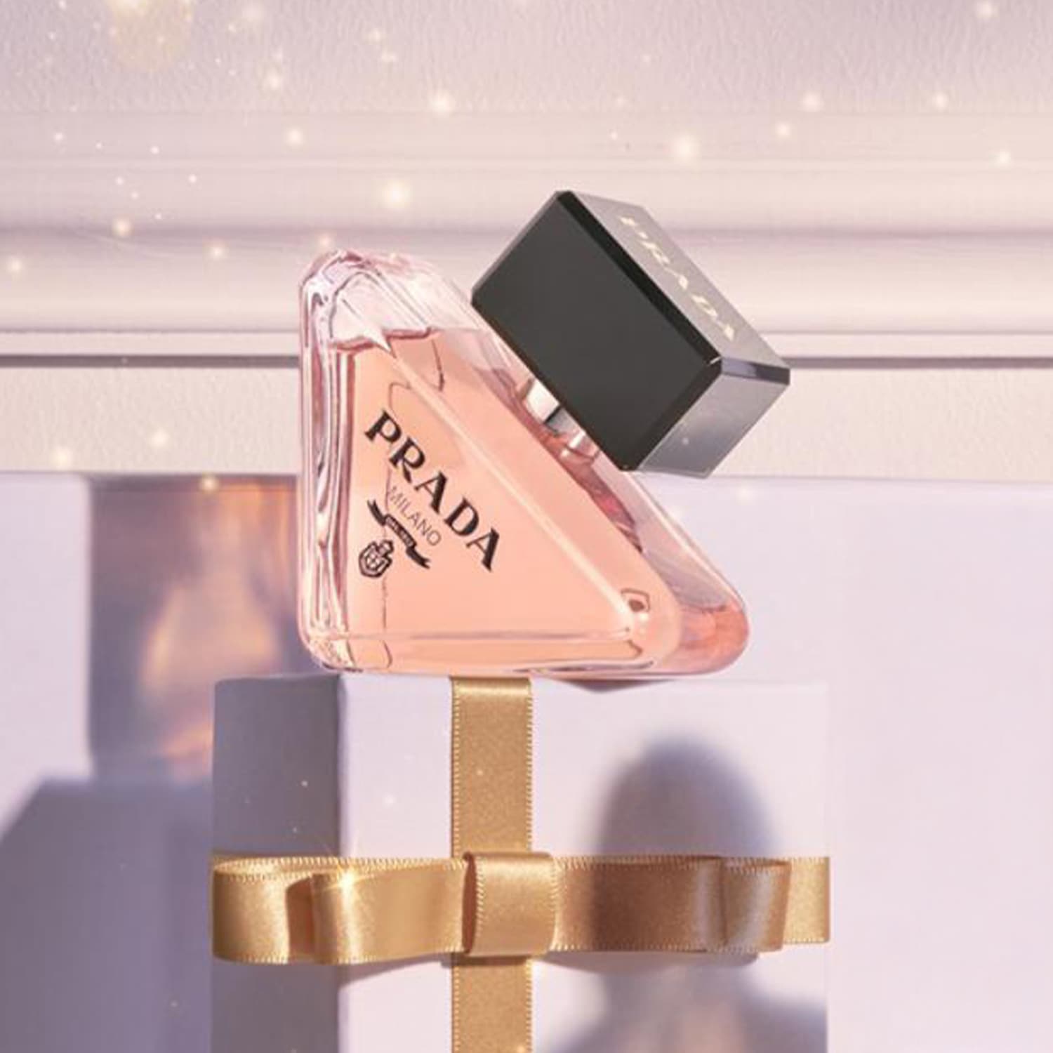 Różowy flakon perfum Prada stoi na białym pudełku przewiązanym złotą wstążką, tworząc elegancką kompozycję.