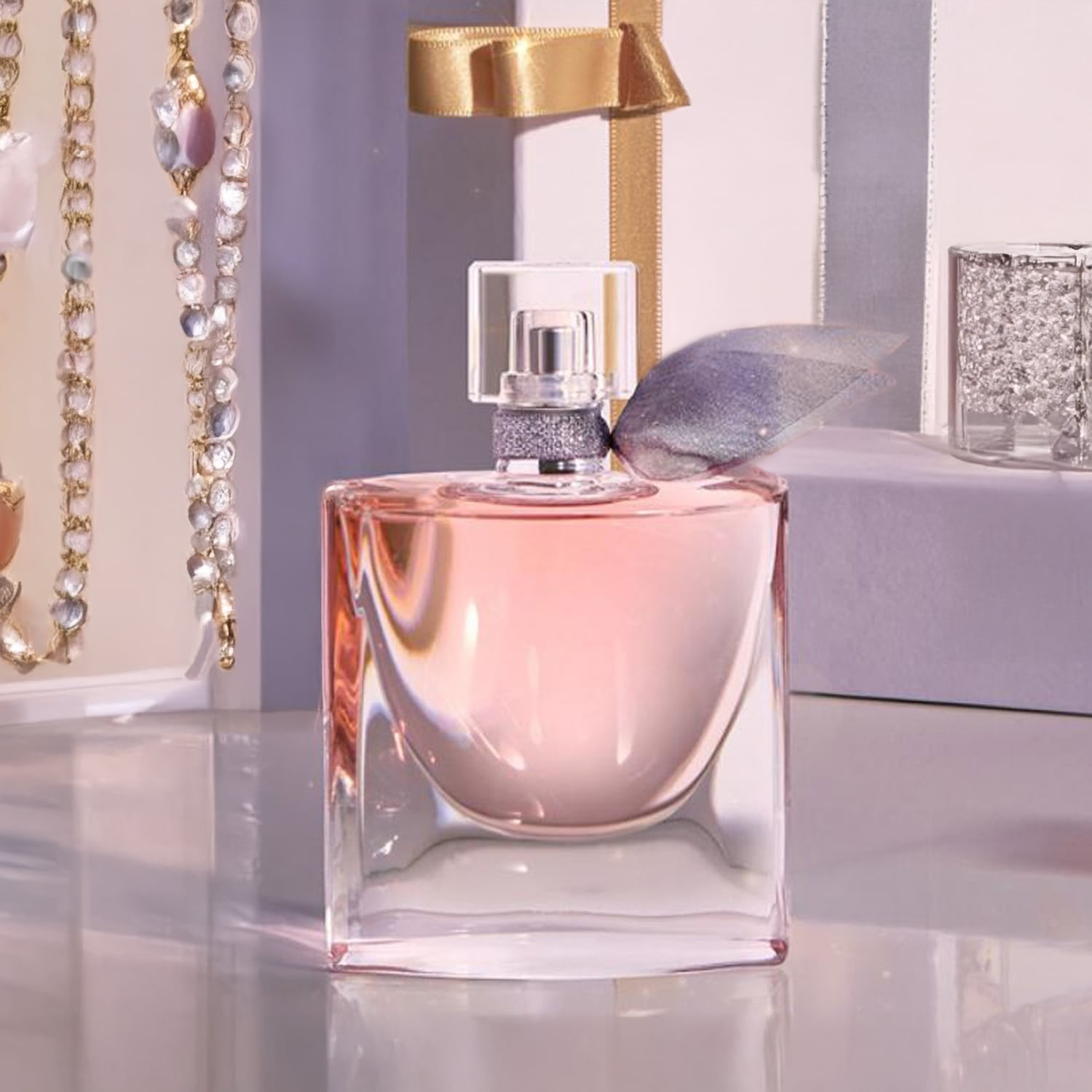 Flakon perfum La Vie Est Belle o pojemności 50 ml z różową zawartością i srebrną kokardką, na błyszczącej powierzchni, w tle ozdobne łańcuszki i pudełka prezentowe.