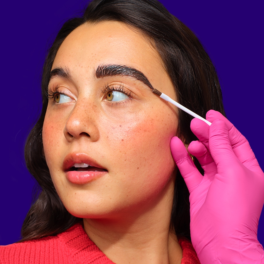 Nahaufnahme einer Frau, die mit einem Pinsel und pinkfarbenen Handschuhen ihre Augenbrauen betont, um einen gepflegten Look zu erzielen.