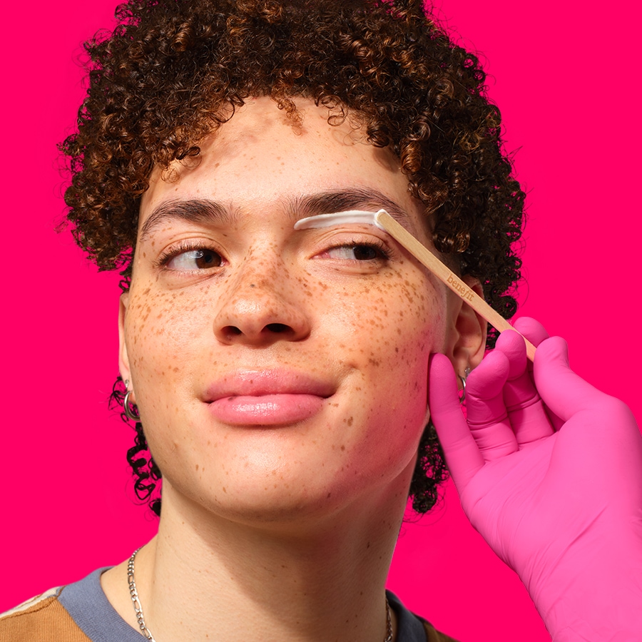 Ein Gesicht mit Sommersprossen, auf dessen Augenbraue mit einem Holzstäbchen weiße Creme aufgetragen wird, während eine Hand mit einem pinkfarbenen Handschuh das Stäbchen hält.