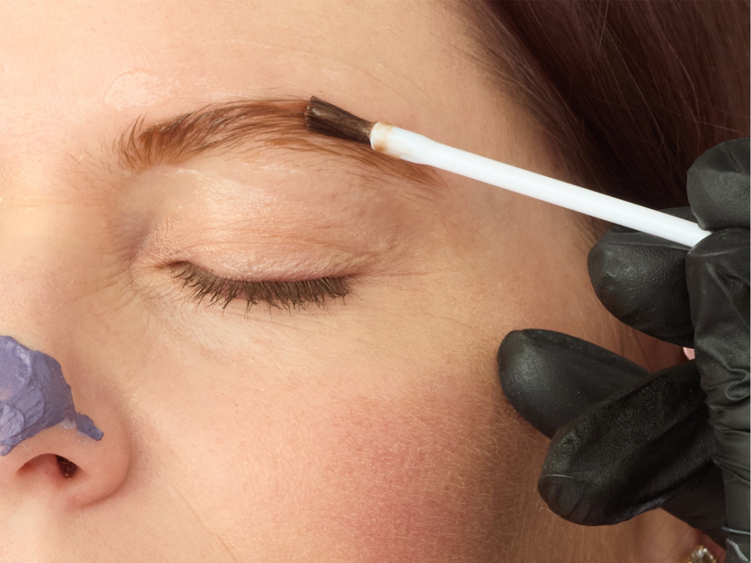 Eine Kosmetikerin mit schwarzen Handschuhen trägt mit einem weißen Pinsel Farbe auf die Augenbrauen einer Frau auf, während sich auf ihrer Nase ein lilafarbenes Produkt befindet.