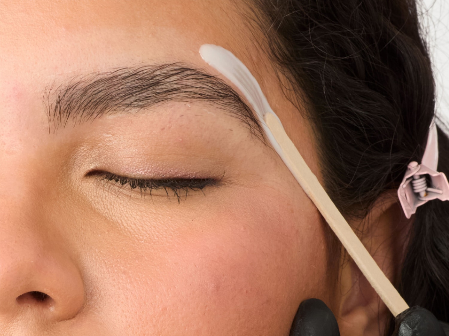 Nahaufnahme einer Frau, deren Augen geschlossen sind, während eine weiße Creme mit einem Holzspatel auf ihre Augenbraue aufgetragen wird.