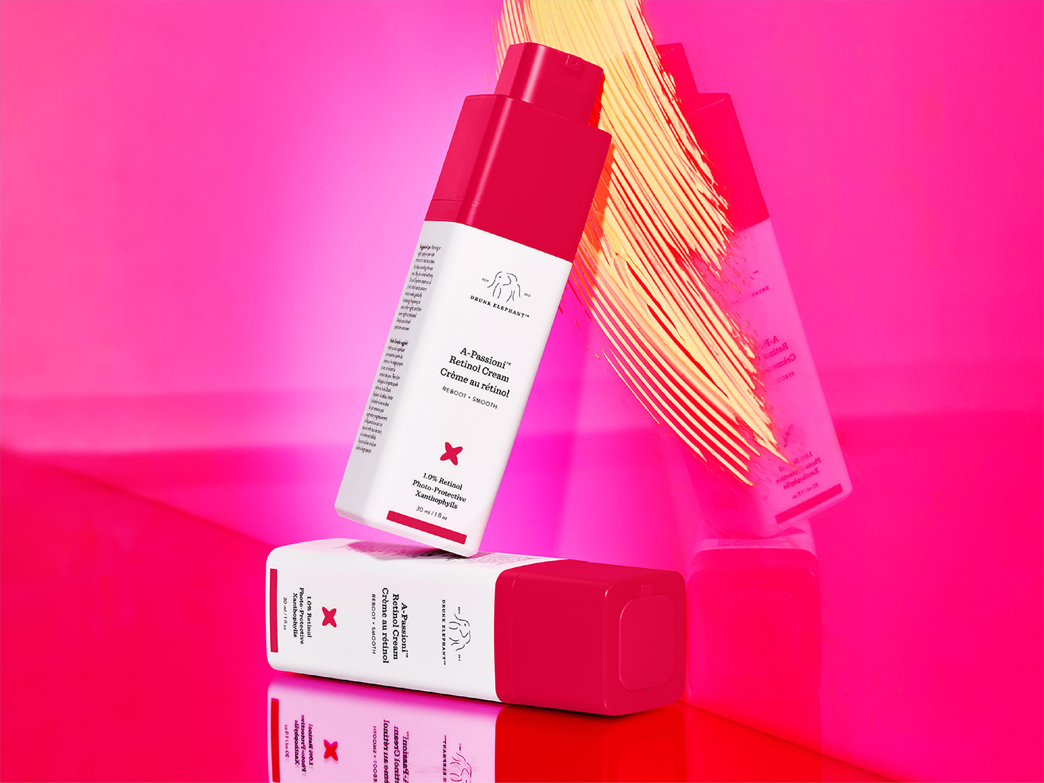 Drei Drunk Elephant A-Passioni Retinol Cremes mit weißer und pinkfarbener Verpackung sind auf pinkfarbenem Hintergrund zu sehen, die Textur ist glatt und cremig.
