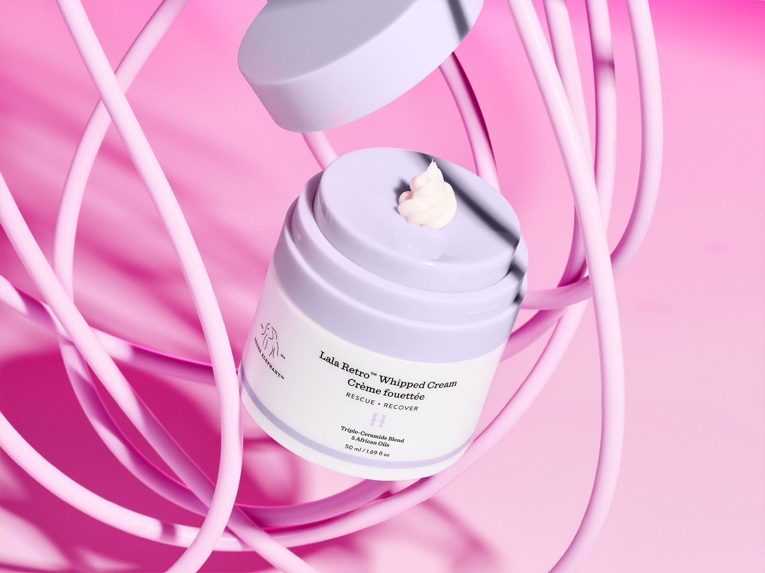 Nahaufnahme von Drunk Elephant Lala Retro Whipped Cream in einer weißen Dose mit lila Deckel, die von rosa Röhren umgeben ist und eine kleine Menge Creme auf dem Deckel zeigt.