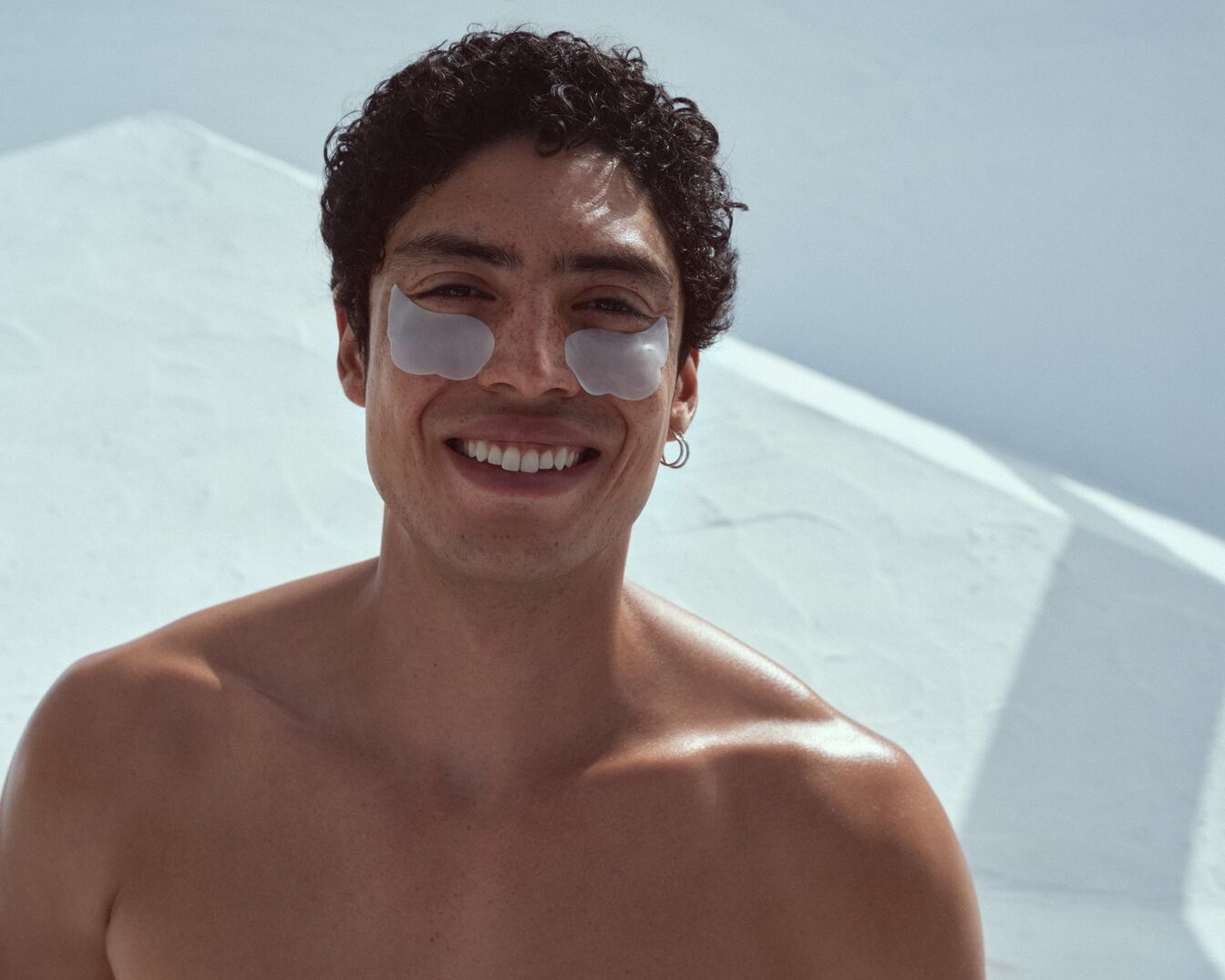 Šypsodamasis jaunas vyras su tamsiais garbanotais plaukais ir sidabriniais auskarais, ant paakių užsidėjęs paakių kaukes.