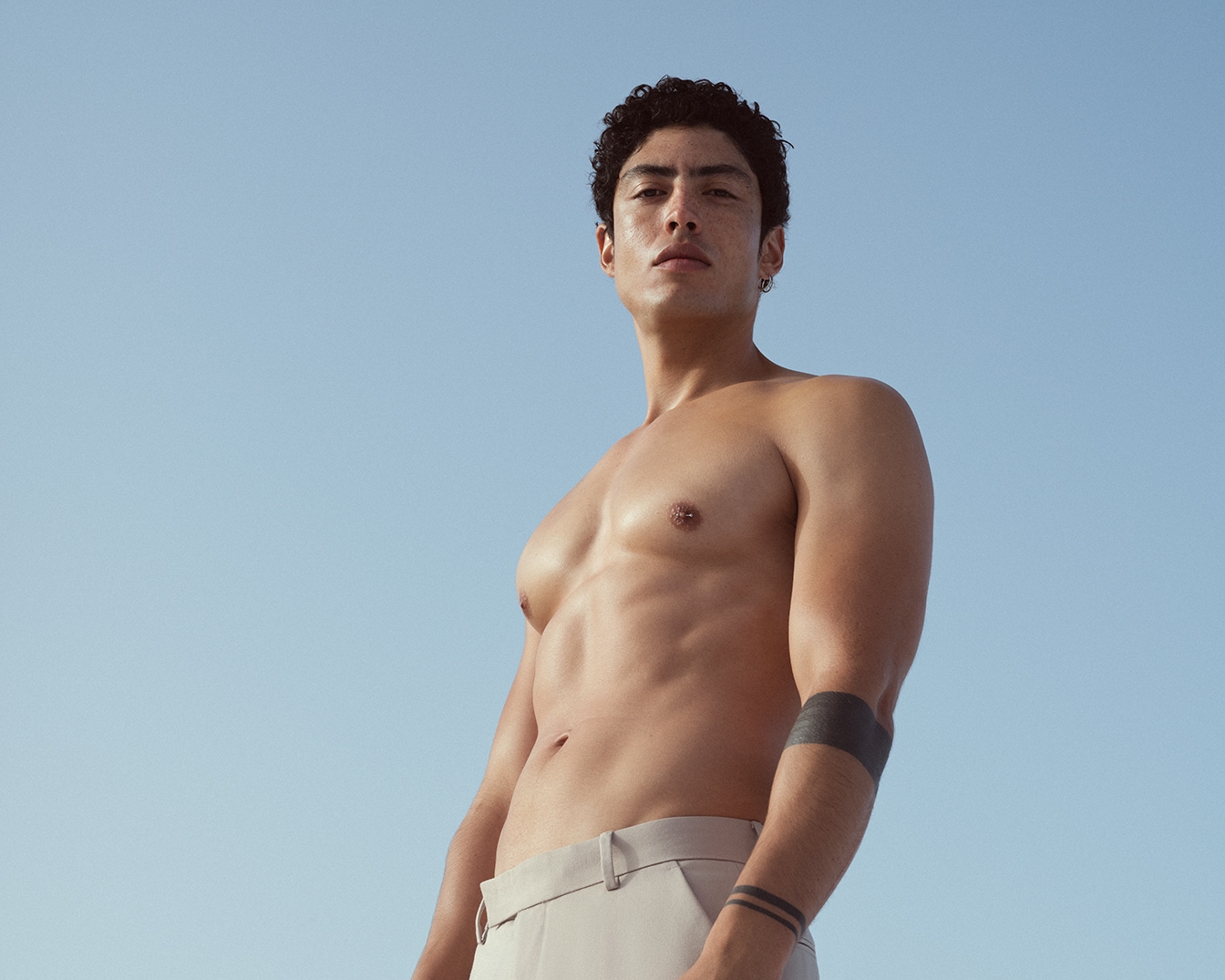 Vyras be marškinių su garbanotais plaukais ir tatuiruotėmis ant rankų, stovintis prieš mėlyną dangų.