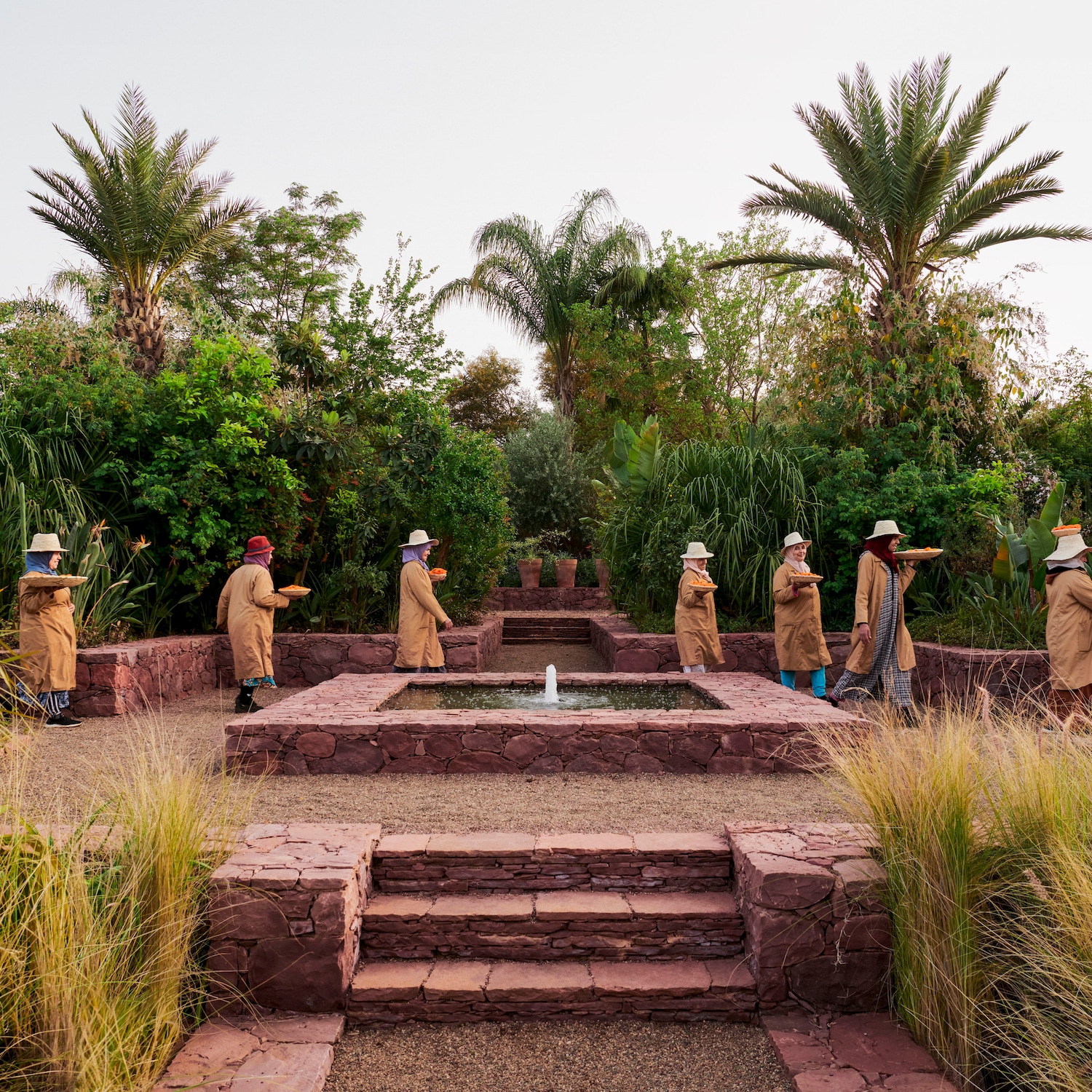 Eine Gruppe von Frauen in beigen Mänteln und Hüten spaziert durch einen üppigen Garten mit einem roten Steinbrunnen und Palmen.