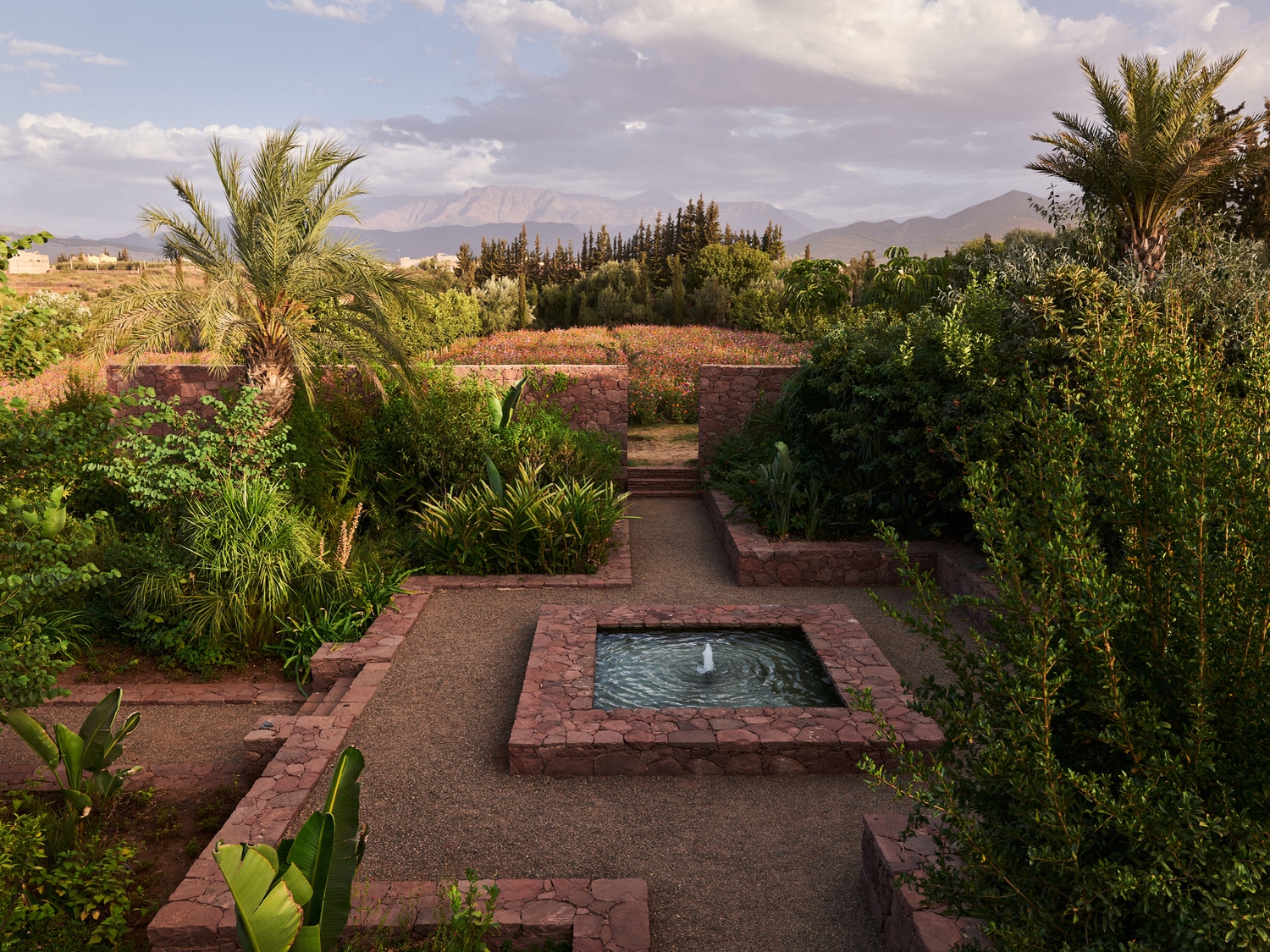 Ein Garten mit einem kleinen Brunnen, umgeben von üppigem Grün und Palmen, mit Bergen im Hintergrund.