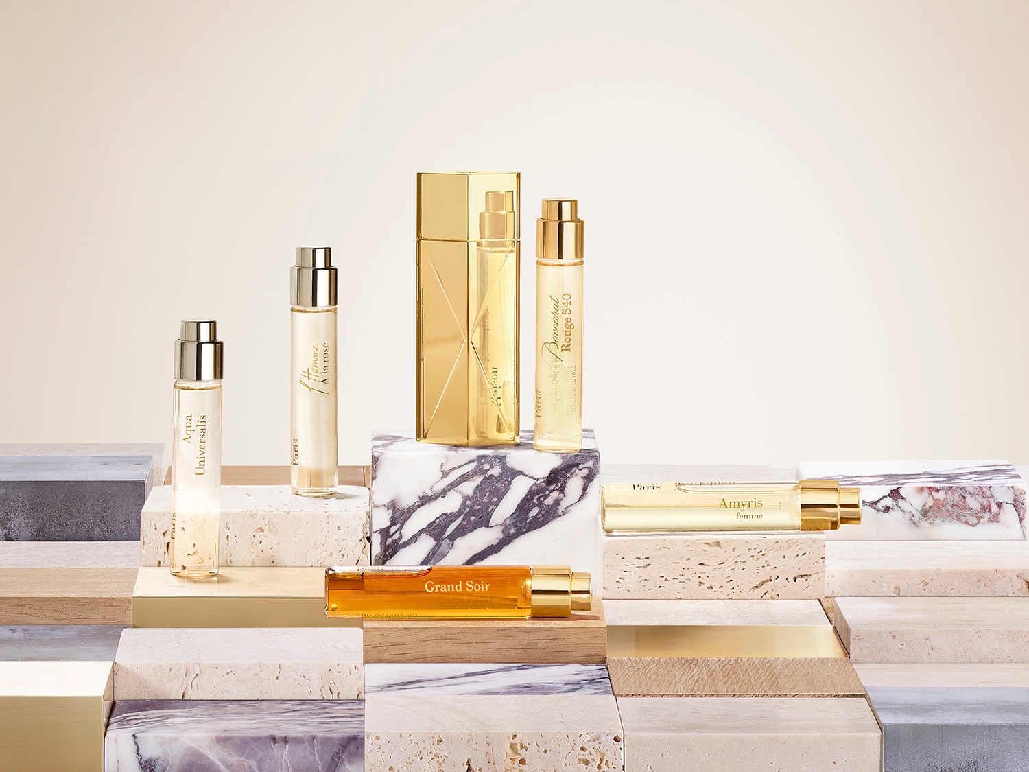 Eine Kollektion goldfarbener Parfümflaschen verschiedener Größen und Formen, darunter Aqua Universalis, A la rose, Grand Soir, Amyris femme und Baccarat Rouge 540, die auf Marmor- und Holzsockeln präsentiert werden.