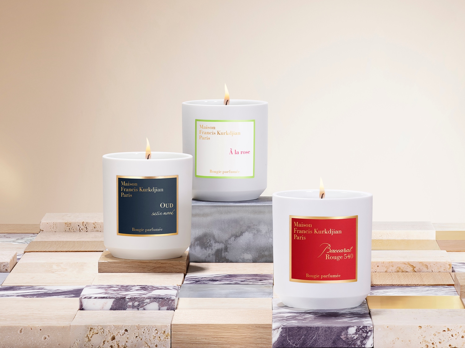Drei weiße Duftkerzen von Maison Francis Kurkdjian Paris, jede mit einem andersfarbigen Etikett: blau für Oud, grün für À la rose und rot für Baccarat Rouge 540.