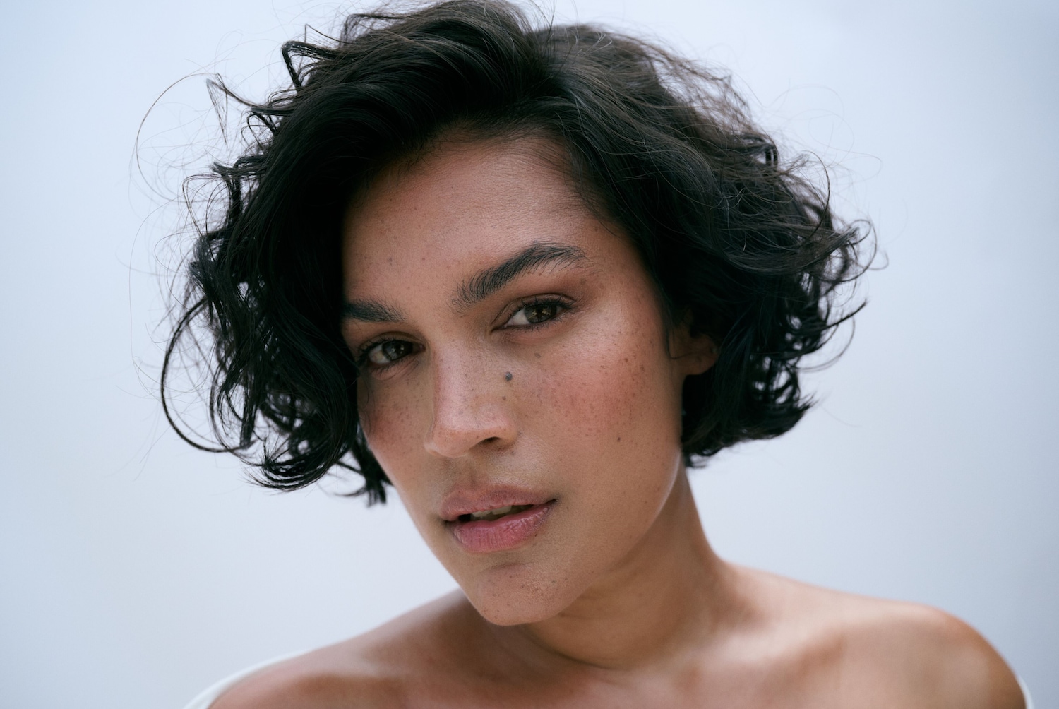 Nahaufnahme einer Frau mit dunklen, lockigen Haaren, Sommersprossen und einem Muttermal im Gesicht, die direkt in die Kamera schaut.