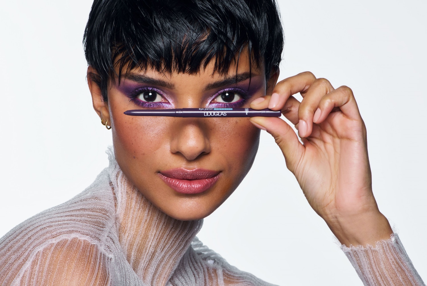 Eine Frau mit lila Lidschatten hält einen violetten Douglas-Eyeliner vor ihre Augen.