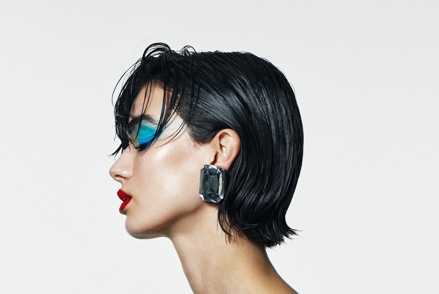 Profil einer Frau mit glänzendem, kurzem, schwarzem Haar, blauem Lidschatten, rotem Lippenstift und einem großen, rechteckigen Ohrring.