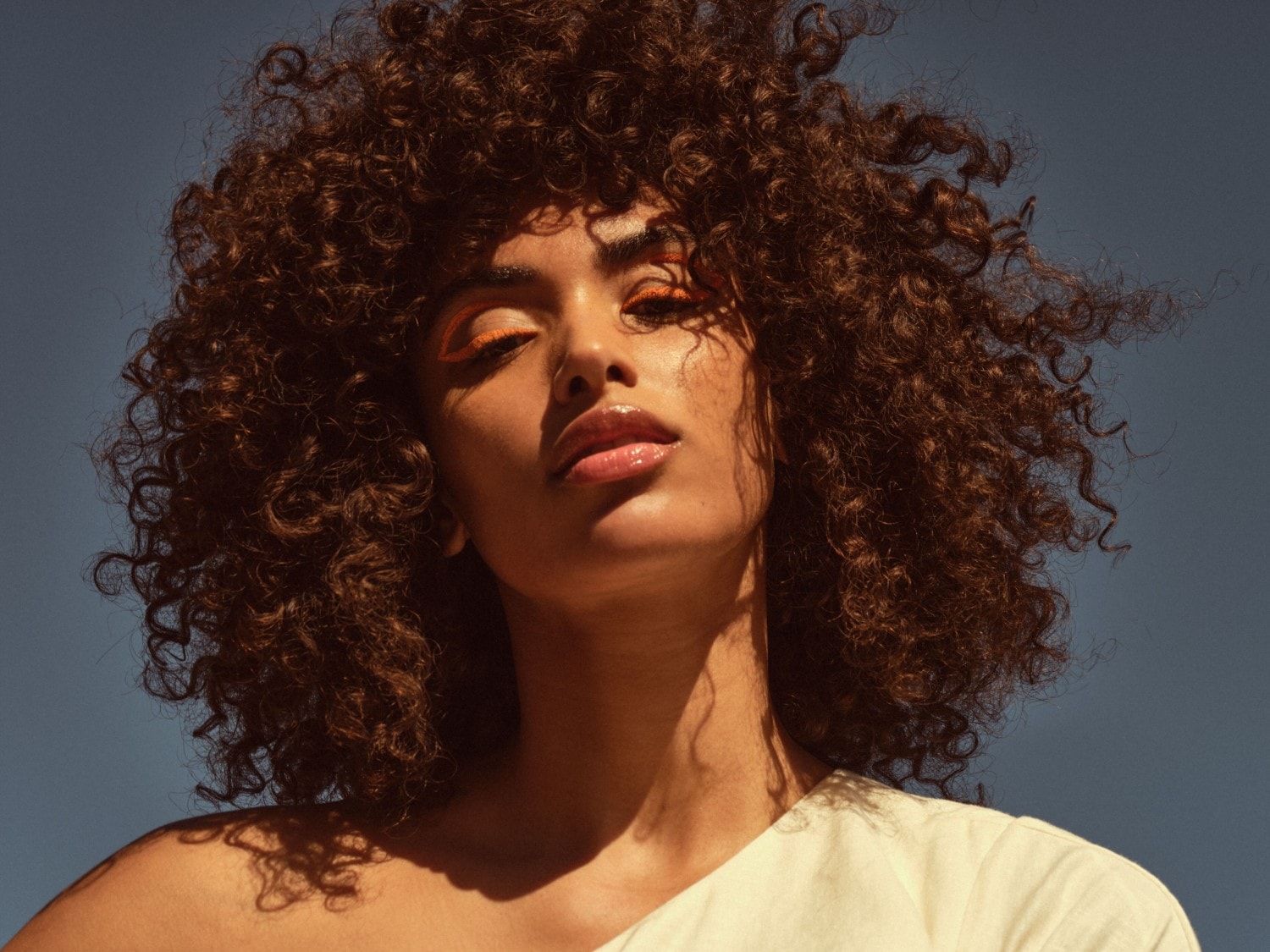 Nahaufnahme einer Frau mit lockigem, braunem Haar, orangefarbenem Eyeliner und glänzenden Lippen vor einem blauen Himmel.