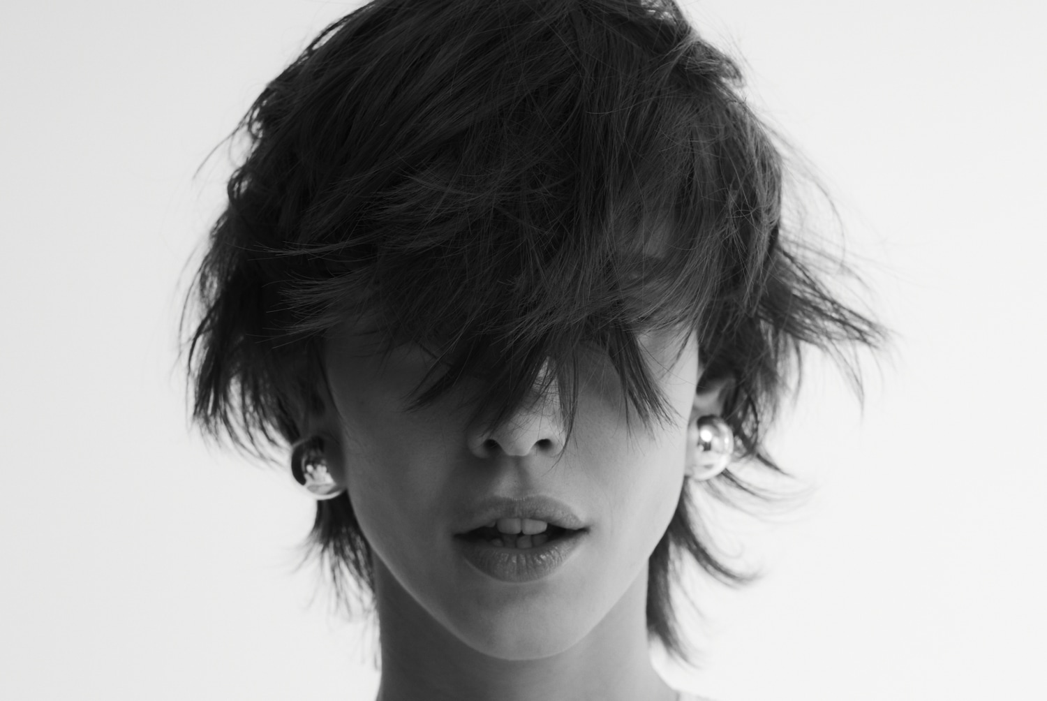 Schwarz-Weiß-Porträt einer Person mit dunklen, zerzausten Haaren, die das Gesicht teilweise bedecken, und silbernen Kugelohrringen.