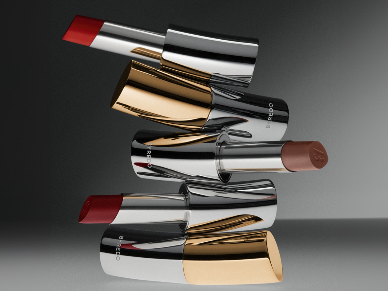 Na obrázku je pyramída rúžov Byredo v odtieňoch červenej a hnedej so striebornými a zlatými obalmi, ktoré vytvárajú luxusný a elegantný vzhľad.