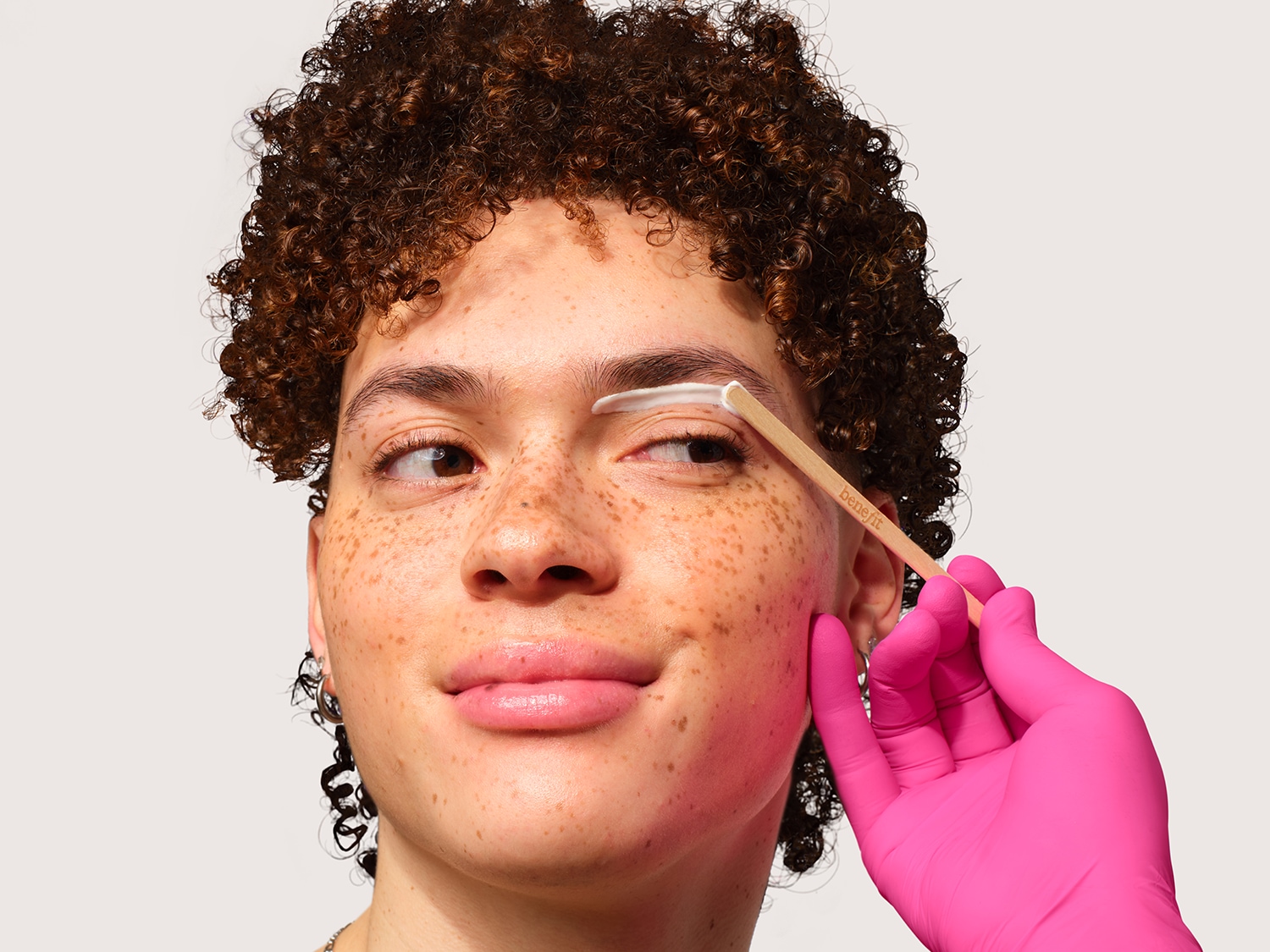 Gros plan d'une personne aux cheveux bouclés se faisant appliquer une crème blanche sur les sourcils avec un bâtonnet par une main gantée de rose, pour une mise en beauté précise et soignée.