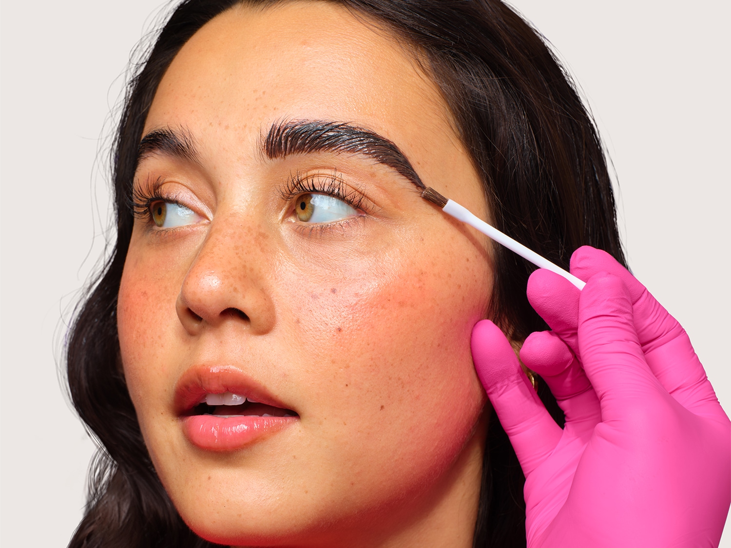 Gros plan d'une femme appliquant un gel à sourcils transparent avec une brosse à bobine blanche, mettant en valeur des sourcils brillants et bien coiffés.