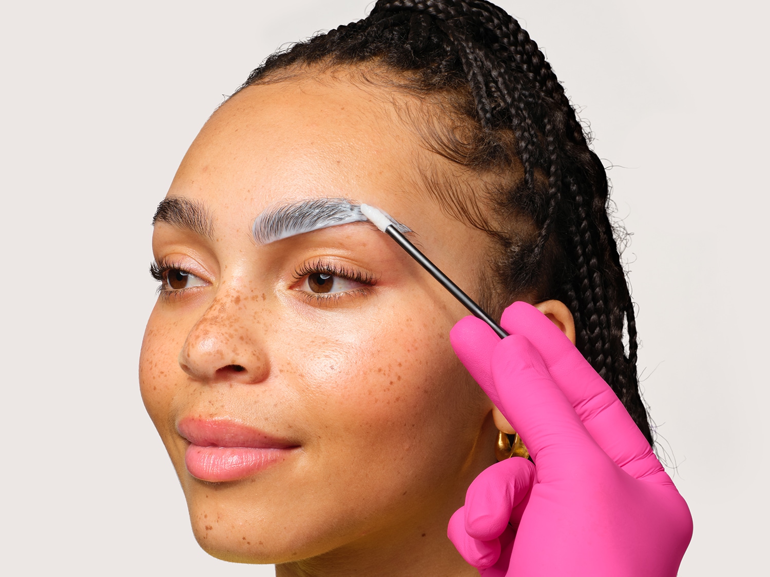 Gros plan d'une femme appliquant un traitement blanc crémeux sur ses sourcils avec un applicateur à pointe fine, portant des gants roses.