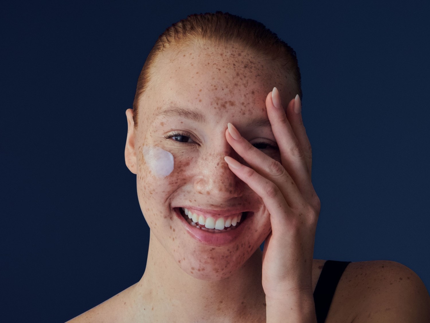 Nahaufnahme einer lächelnden Frau mit Sommersprossen und einem Klecks weißer Creme auf der Wange, die ihr Gesicht mit der Hand berührt.