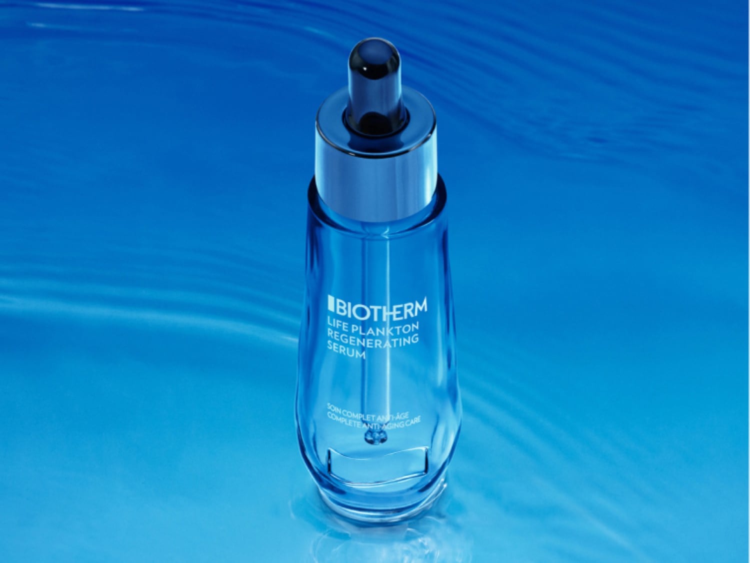 Das Biotherm Life Plankton Regenerating Serum in einer durchsichtigen, blau getönten Glasflasche mit Pipette, die auf einer glatten, blauen Oberfläche steht.