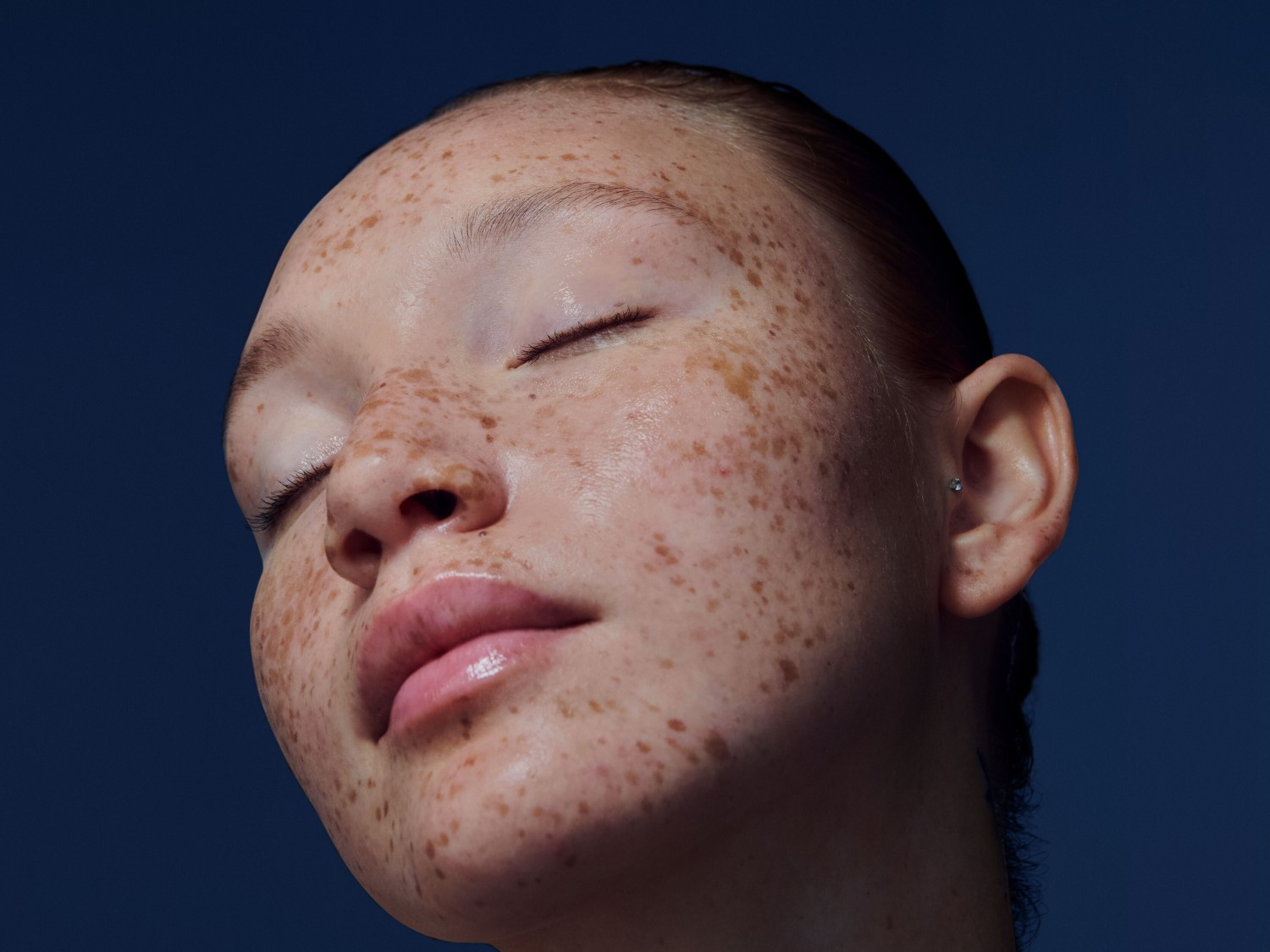 Nahaufnahme eines jungen, hellhäutigen Mädchens mit Sommersprossen, geschlossenen Augen und glänzenden Lippen vor einem dunkelblauen Hintergrund.