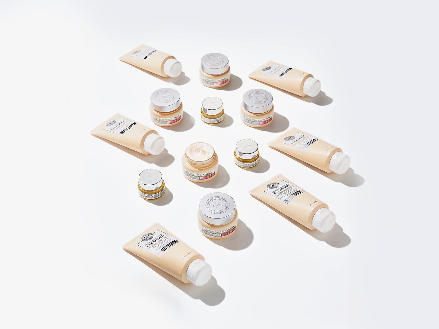Különféle IT Cosmetics termékek láthatók, beleértve a Confidence in a Cream tégelyeket és a krém színű tisztító tubusokat, amelyek egy fehér felületen vannak elrendezve.