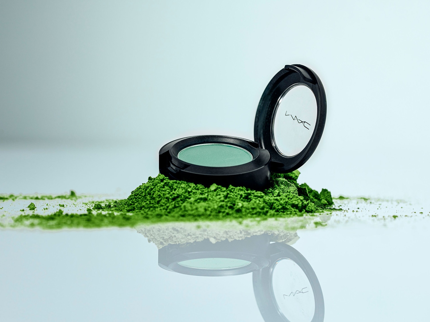 Otwarty, okrągły, czarny puder do powiek MAC z zielonym cieniem w środku, leżący na kopcu zielonego proszku, odbijający się na błyszczącej powierzchni.