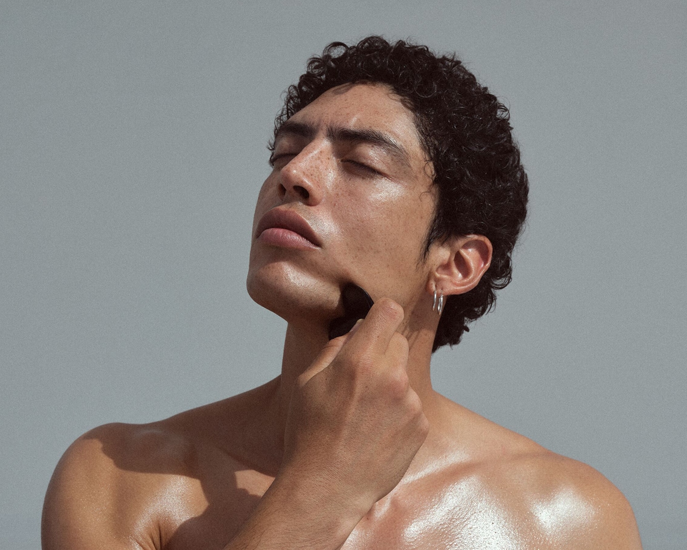 Een man met krullend haar en een zilveren oorbel masseert zijn gezicht met een zwarte gezichtsroller tegen een grijze achtergrond.