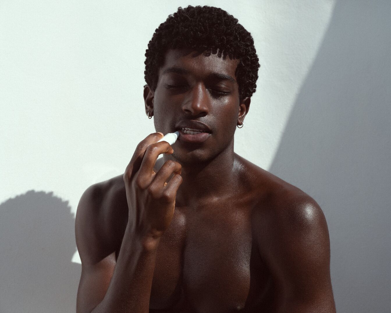 Een getinte man met kort krullend haar brengt een witte lippenbalsem aan op zijn lippen tegen een witte achtergrond.