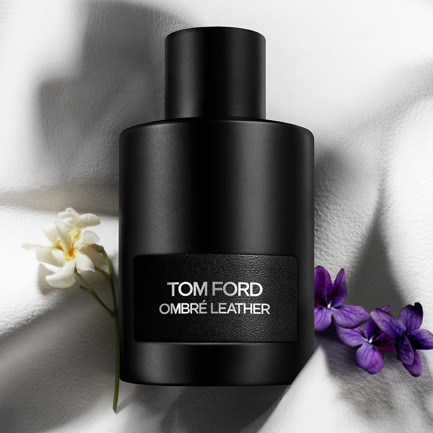 Découvrez le parfum Tom Ford Ombré Leather dans son flacon noir élégant, agrémenté de touches florales blanches et violettes.