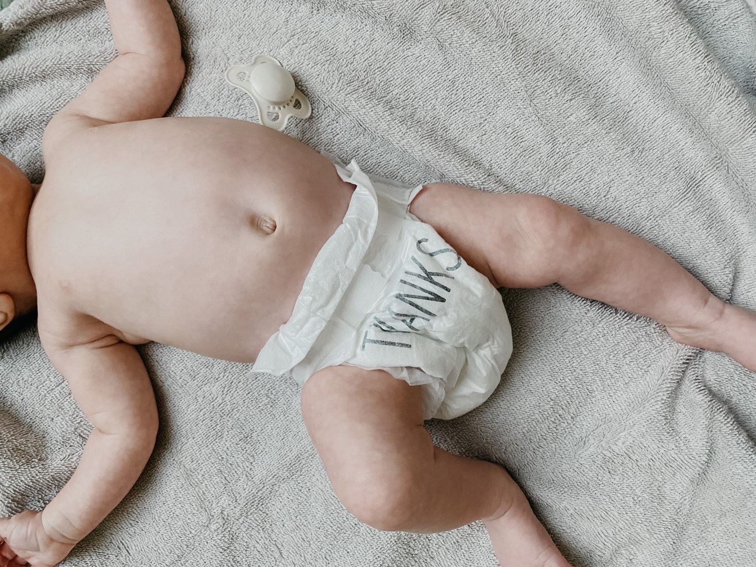 Draufsicht auf ein Baby mit Windel, auf der das Wort 'THANKS' steht, daneben ein Schnuller auf einem grauen Tuch.